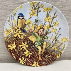 Può includere: Un piatto decorativo con un passero azzurro e una fata seduti in un nido di fiori gialli. Il piatto è bianco con uno sfondo blu chiaro.