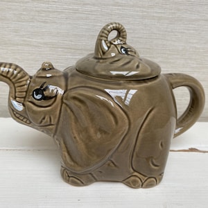 Könnte beinhalten: Eine braune Teekanne aus Keramik in Form eines Elefanten mit Deckel und Ausguss. Die Teekanne hat bemalte Details, darunter Augen und einen Rüssel.