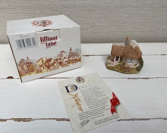 Może przedstawiać: Miniaturowy domek z dachem z trzciny, kamiennymi ścianami i małym ogrodem. Domek jest częścią kolekcji o nazwie "Lilliput Lane" i jest dostarczany z aktem własności. Akt jest wydrukowany na białej kartce z czerwoną pieczęcią woskową.