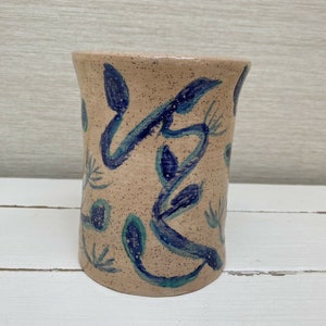 Könnte beinhalten: Eine beige Keramikvase mit einem blauen und grünen Blumendesign. Die Vase hat eine strukturierte Oberfläche und eine zylindrische Form.
