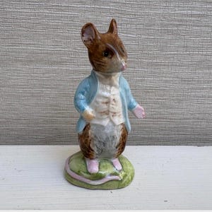 May include: A ceramic mouse figurine wearing a light blue jacket and white waistcoat. The mouse stands on a green base with pink detailing. The figurine has brown fur and pink hands and feet.