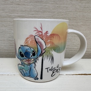 May include: White ceramic mug featuring a smiling blue cartoon character with large eyes. The mug has a tropical sunset design with palm trees and the text "Take it Easy". The mug has a white handle.