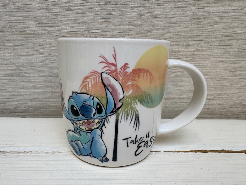 May include: White ceramic mug featuring a smiling blue cartoon character with large eyes. The mug has a tropical sunset design with palm trees and the text "Take it Easy". The mug has a white handle.