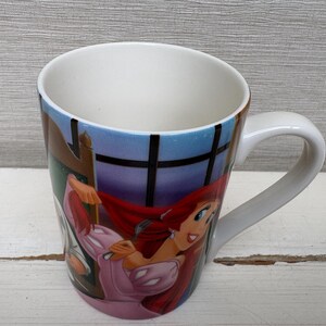 May include: White ceramic mug featuring a colorful illustration of a cartoon character with red hair and a pink dress. The mug has a curved handle and a blue and white background.