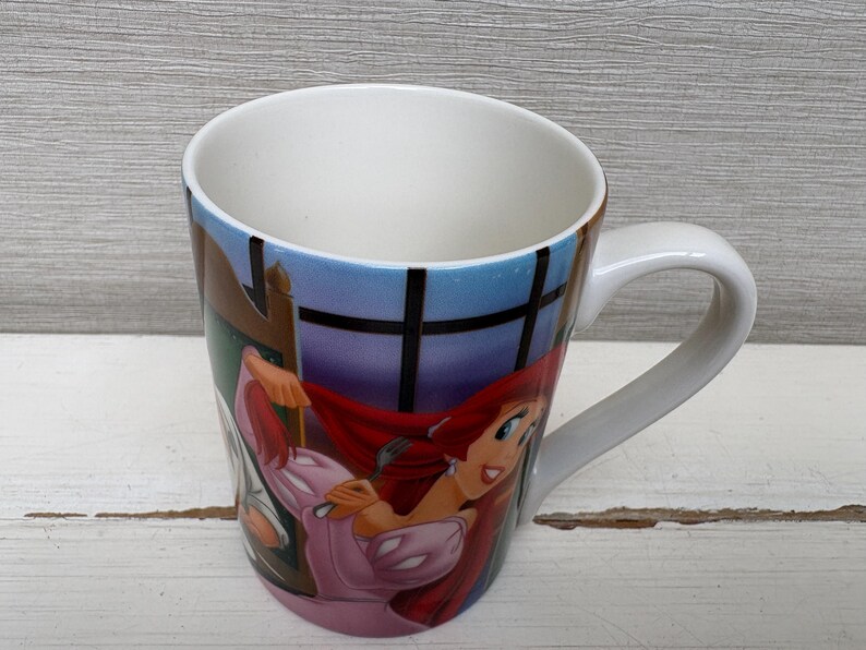 May include: White ceramic mug featuring a colorful illustration of a cartoon character with red hair and a pink dress. The mug has a curved handle and a blue and white background.