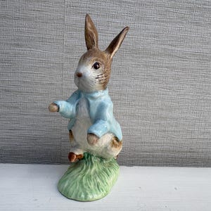 Könnte beinhalten: Eine Keramikfigur eines Kaninchens, das auf einem grünen Grasuntergrund steht. Das Kaninchen hat braunes und weißes Fell, dunkle Augen und trägt eine hellblaue Jacke. Die Figur ist in einer aufrechten, wachsamen Haltung dargestellt.