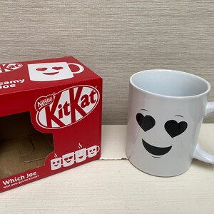 May include: A white ceramic mug with a black smiley face and heart-shaped eyes. The mug is sitting on a white surface next to a red cardboard box with the words "Nestle KitKat" and "Dreamy Joe" printed on it.