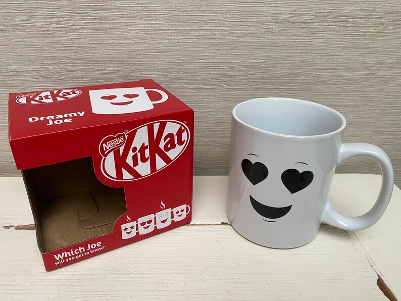 May include: A white ceramic mug with a black smiley face and heart-shaped eyes. The mug is sitting on a white surface next to a red cardboard box with the words "Nestle KitKat" and "Dreamy Joe" printed on it.