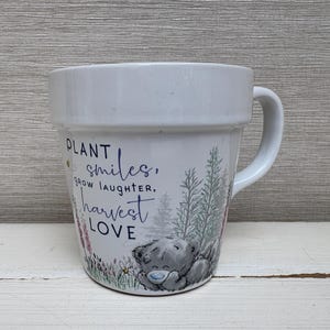 Może przedstawiać: Biały ceramiczny kubek z uchwytem, z napisem "Plant smiles, grow laughter, harvest love". Kubek ma wzór szarego misia, kwiatów i drzew. Kubek nadaje się do gorących lub zimnych napojów.