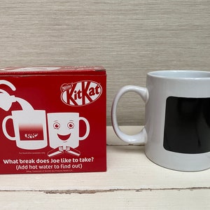 May include: A red box with a white Kit Kat logo and a white mug with a black square on the front. The box has a picture of two mugs, one with a Kit Kat logo and the other with a smiling face. The text on the box reads "What break does Joe like to take? (Add hot water to find out)"