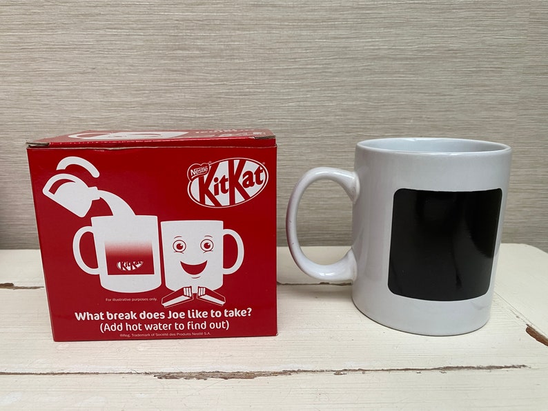 May include: A red box with a white Kit Kat logo and a white mug with a black square on the front. The box has a picture of two mugs, one with a Kit Kat logo and the other with a smiling face. The text on the box reads "What break does Joe like to take? (Add hot water to find out)"