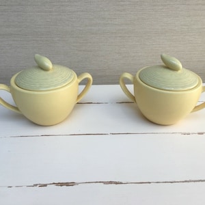 Könnte beinhalten: Zwei gelbe Zuckerdosen aus Keramik mit Deckeln. Die Deckel haben ein hellgrünes Spiralmuster und einen kleinen, blattartigen Knauf.