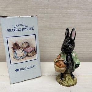 Puede incluir: Una figura de cerámica de un conejo negro con una chaqueta verde claro y una cesta tejida, sobre una base texturizada. A su lado hay una caja de cartón azul claro con la inscripción The World of Beatrix Potter y Royal Albert.