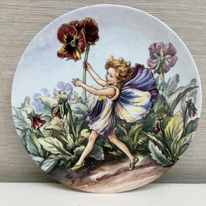 Könnte beinhalten: Dekorative Platte mit einer skurrilen Illustration einer Fee in einem Garten. Die Fee mit lila und gelben Flügeln greift nach einer großen, dunkelroten Blume. Die Platte hat einen hellblauen Hintergrund mit anderen Blumen und Blättern.