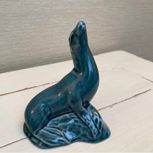 Puede incluir: Una figura de foca de cerámica de color verde azulado. La foca está representada en posición vertical, con la cabeza y el cuello extendidos. La figura tiene un acabado brillante y descansa sobre una base texturizada, que sugiere agua. El diseño es suave y detallado.