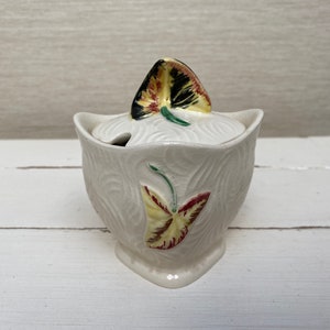 May include: White ceramic sugar bowl with a lid featuring a yellow, red, and black leaf design. The bowl has a textured surface and a leaf design on the front.