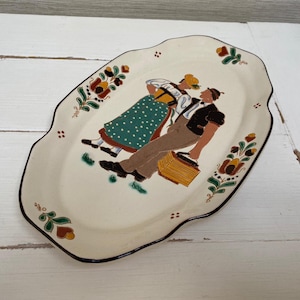 Może przedstawiać: Biała owalna ceramiczna taca do serwowania z brązową krawędzią i malowanym wzorem dwóch osób w tradycyjnych strojach. Wzór zawiera zielone, brązowe i czerwone akcenty kwiatowe.