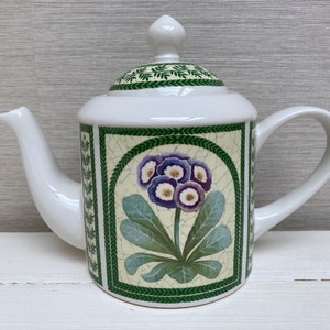 Könnte beinhalten: Weiße Teekanne aus Keramik mit einem grün-weißen Blumendesign. Die Teekanne hat einen Deckel mit einem Knopf oben und einen Ausguss auf einer Seite. Das Blumendesign zeigt eine Gruppe von lila und weißen Blüten mit grünen Blättern.