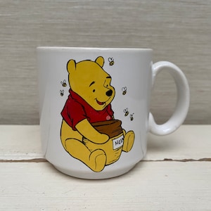 Könnte beinhalten: Weiße Keramiktasse mit einer Illustration von Winnie Puuh. Puuh hält einen Honigtopf und ist von Bienen umgeben. Die Tasse hat einen Henkel.