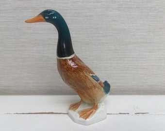 May include: A ceramic mallard duck figurine featuring a glossy finish with a dark green head, a bright orange beak, and a brown feathered body. The duck stands on a white base, with its neck extended forward in a realistic, alert posture.