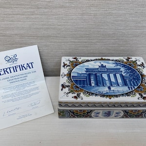 Könnte beinhalten: Eine blaue und weiße Keramikdose mit floralem Design und einem Bild des Brandenburger Tors. Die Dose ist mit "200 Jahre Brandenburger Tor Gedenkausgabe Fayence-Dose" beschriftet.
