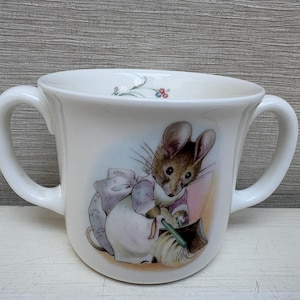 Peut inclure: Tasse à bébé en céramique blanche avec deux anses. La tasse présente une charmante illustration d'une souris vêtue d'une tenue rose et violette. La souris tient un balai. La tasse a un motif floral sur le bord.