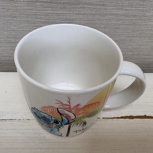 May include: White ceramic mug featuring a colorful illustration of Stitch from Lilo & Stitch. The design includes Stitch, palm trees, and a sunset. The mug has a curved shape and a white handle.
