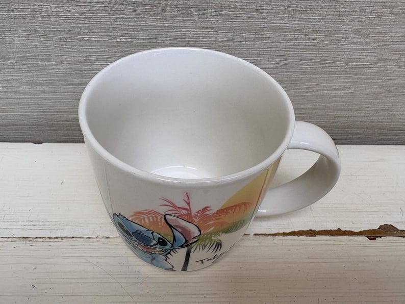 May include: White ceramic mug featuring a colorful illustration of Stitch from Lilo & Stitch. The design includes Stitch, palm trees, and a sunset. The mug has a curved shape and a white handle.