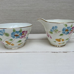 Könnte beinhalten: Ein weißes Zucker- und Milchkännchen-Set aus Keramik mit einem Blumendesign. Das Design zeigt blaue, rosa und gelbe Blumen auf weißem Hintergrund.
