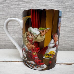 May include: A ceramic coffee mug with a white handle and a colorful cartoon illustration. The illustration features a chef holding a plate of food, a red lobster, and other characters from a popular animated film.