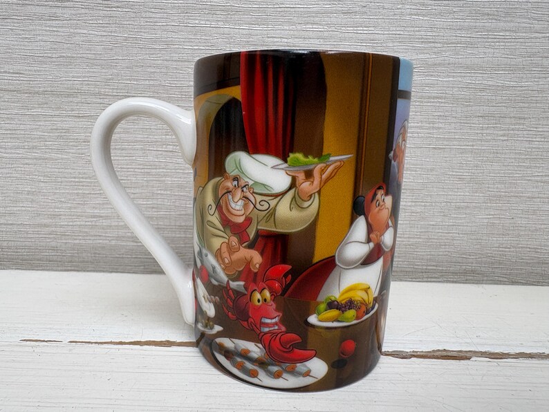 May include: A ceramic coffee mug with a white handle and a colorful cartoon illustration. The illustration features a chef holding a plate of food, a red lobster, and other characters from a popular animated film.