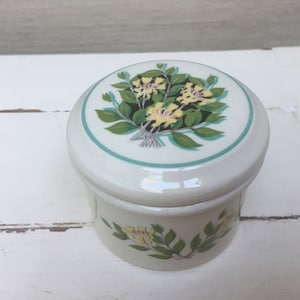 Könnte beinhalten: Eine weiße Keramik-Schmuckdose mit floralem Design. Der Deckel zeigt einen Strauß gelber Blumen mit grünen Blättern auf weißem Hintergrund mit hellblauem Rand. Die Unterseite der Dose hat einen einzelnen Zweig mit gelben Blumen und grünen Blättern.