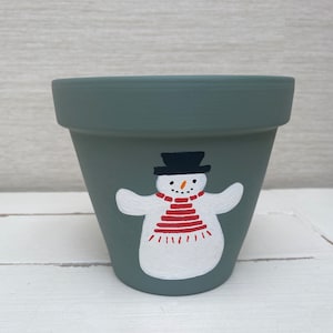 Könnte beinhalten: Ein grüner Terrakotta-Blumentopf mit handgemaltem Schneemann-Design. Der Schneemann trägt einen schwarzen Hut, einen rot-weißen Schal und hat einen weißen Körper mit schwarzen Augen und einer Karottennase.