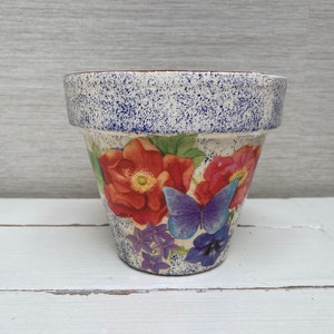 Könnte beinhalten: Ein weißer Keramik-Blumentopf mit blauer, gesprenkelter Glasur. Der Topf ist mit einem Blumendesign verziert, das rote, rosa und violette Blumen und einen blauen Schmetterling zeigt.