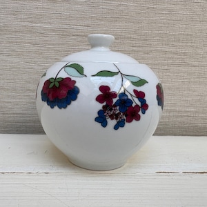 May include: A white ceramic sugar bowl with a rounded body and a matching lid. The exterior features a decorative pattern of stylised flowers in shades of deep magenta, blue, and green, with thin green stems and leaves encircling the centre.