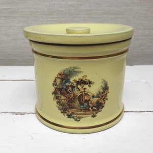 Könnte beinhalten: Eine gelbe Keramikdose mit Deckel, verziert mit einem Vintage-Blumendesign mit einer Frau und einem Kind in einem Garten.