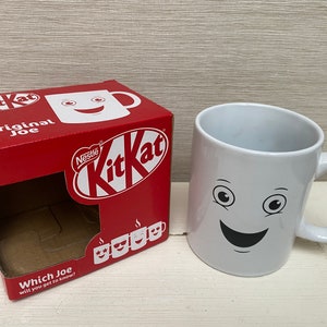 May include: A white ceramic mug with a smiling face and two eyes. The mug is next to a red box with the words "Nestle KitKat Original Joe" and a picture of a KitKat bar.