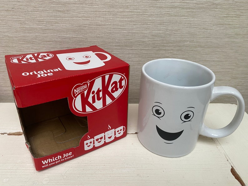 May include: A white ceramic mug with a smiling face and two eyes. The mug is next to a red box with the words "Nestle KitKat Original Joe" and a picture of a KitKat bar.