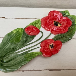 Könnte beinhalten: Eine Keramik-Servierplatte mit Mohnblumen-Design. Die Platte ist weiß mit grünen Blättern und roten Mohnblumen. Die Blüten haben schwarze Zentren. Die Platte ist etwa 30 cm lang.