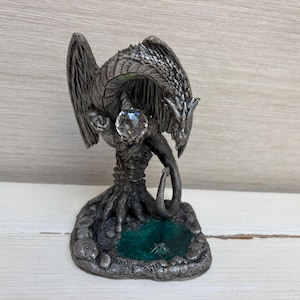 Könnte beinhalten: Eine silberfarbene Drachenfigur aus Metall auf einem strukturierten Sockel mit einem kleinen türkisfarbenen Becken. Der Drache hat detaillierte Schuppen, große Flügel und hält eine klare, facettierte Kristallkugel zwischen seinen Vorderkrallen.