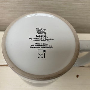 May include: White ceramic mug with a black Nestle logo and instructions for care. The mug is not dishwasher or microwave safe. The text reads "Nestle. Reg. Trademark of Societe des Produits Nestle S.A. Made in China. Not suitable for use in a Dishwasher or Microwave."