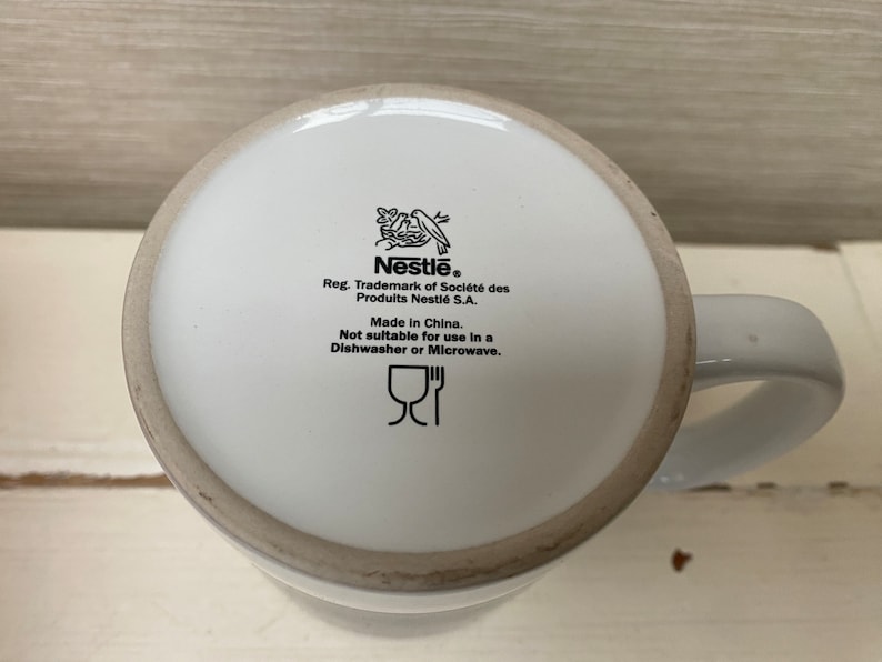 May include: White ceramic mug with a black Nestle logo and instructions for care. The mug is not dishwasher or microwave safe. The text reads "Nestle. Reg. Trademark of Societe des Produits Nestle S.A. Made in China. Not suitable for use in a Dishwasher or Microwave."