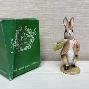 May include: A ceramic figurine of a rabbit wearing a brown jacket and holding a green bag, standing next to a green John Beswick box with white text that reads John Beswick Since 1894 and Hand-made Ceramic Sculpture.