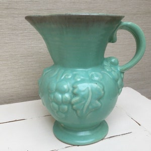 Könnte beinhalten: Eine hellgrüne Keramikkanne mit Henkel und Traubenranken-Design. Die Kanne hat eine glasierte Oberfläche und einen leicht dunkleren grünen Rand.