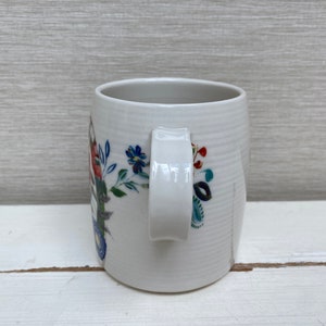 以下が含まれることがあります： 青、赤、緑の色合いの花柄が描かれた白いセラミック製のマグカップ。マグカップは持ち手があり、わずかに丸みを帯びた形をしています。