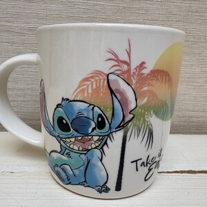 May include: White ceramic mug featuring the character Stitch from Lilo & Stitch. The mug has a curved handle and a colorful design of Stitch, palm trees, and a sunset. The text "Take it Easy" is also visible.