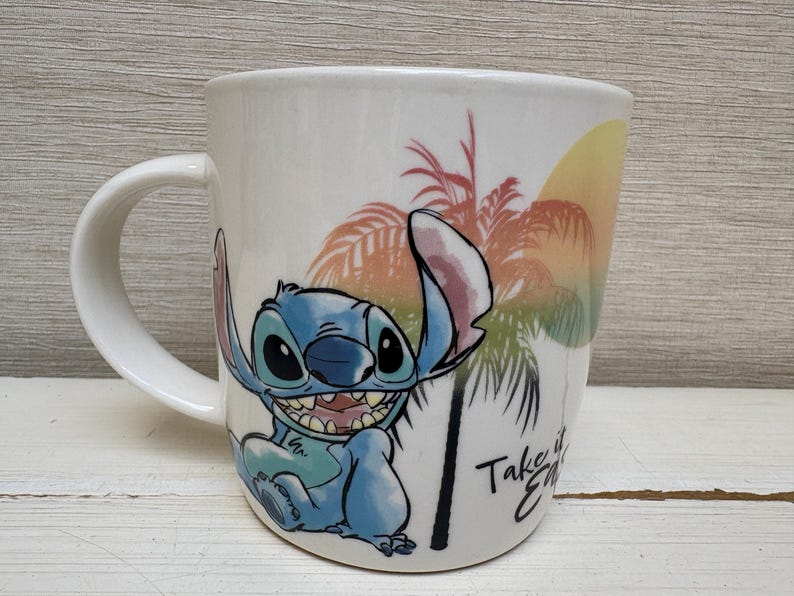 May include: White ceramic mug featuring the character Stitch from Lilo & Stitch. The mug has a curved handle and a colorful design of Stitch, palm trees, and a sunset. The text "Take it Easy" is also visible.