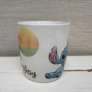May include: White ceramic mug featuring a cartoon illustration of Stitch from Lilo & Stitch. The mug has a colorful design with Stitch and the words "Make it Easy". The mug is cylindrical and has a smooth surface.