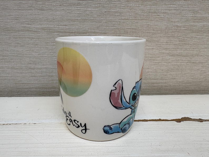 May include: White ceramic mug featuring a cartoon illustration of Stitch from Lilo & Stitch. The mug has a colorful design with Stitch and the words "Make it Easy". The mug is cylindrical and has a smooth surface.
