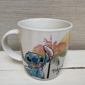 May include: White ceramic mug featuring a cartoon image of Stitch from Lilo & Stitch. The mug has a curved handle and a colorful design of Stitch, palm trees, and a sunset. The text "Take it Easy" is also visible.